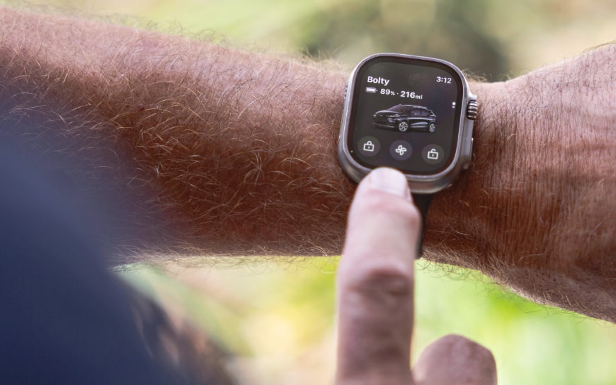 A hand taps a smartwatch screen displaying the myChevrolet app, showing remote controls for a Bolt car.