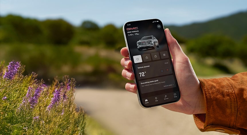 A person's hand holds a black smartphone displaying the GMC mobile app showing the vehicle's remote start and climate control features against a blurred background in an outdoor setting.
