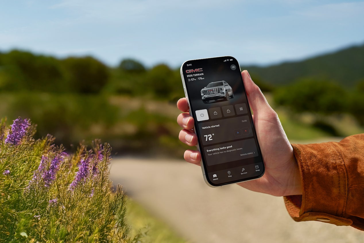 A person's hand holds a black smartphone displaying the GMC mobile app showing the vehicle's remote start and climate control features against a blurred background in an outdoor setting.