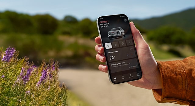 A person's hand holds a black smartphone displaying the GMC mobile app showing the vehicle's remote start and climate control features against a blurred background in an outdoor setting.