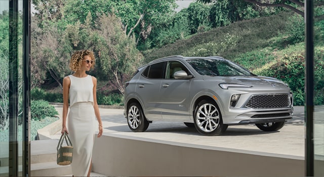 A woman walking into her house with her Buick vehicle parked in the driveway