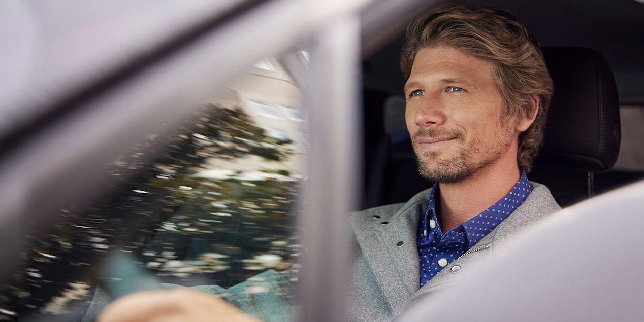 A smiling man with blonde hair and a beard drives a car.