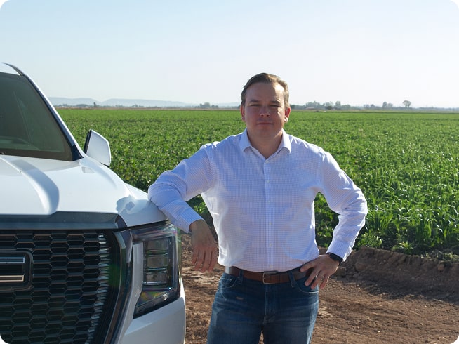 A smiling man, Milas R., in a white button-down shirt is standing in a field of green crops, leaning against the front of his white GMC Yukon with hands-free driver assistance technology from OnStar.