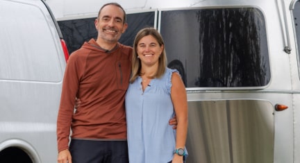 A man and a woman standing next to each other in front of their van and camper.