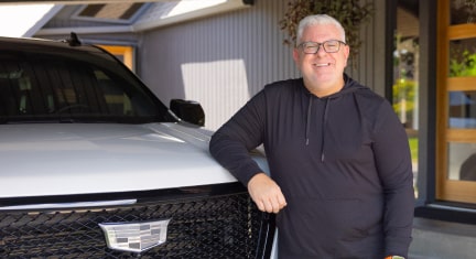  Jason Cupp, a middle-aged man wearing a black hoodie, stands next to the grille of a white Cadillac parked in a garage.