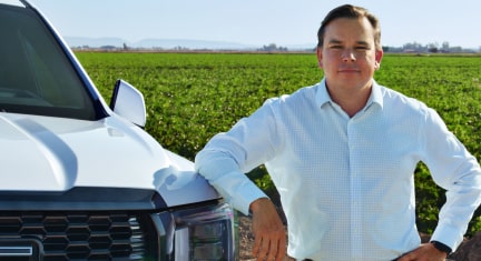 A smiling man, Milas R., in a white button-down shirt is standing in a field of green crops, leaning against the front of his white GMC Yukon with hands-free driver assistance technology from OnStar.
