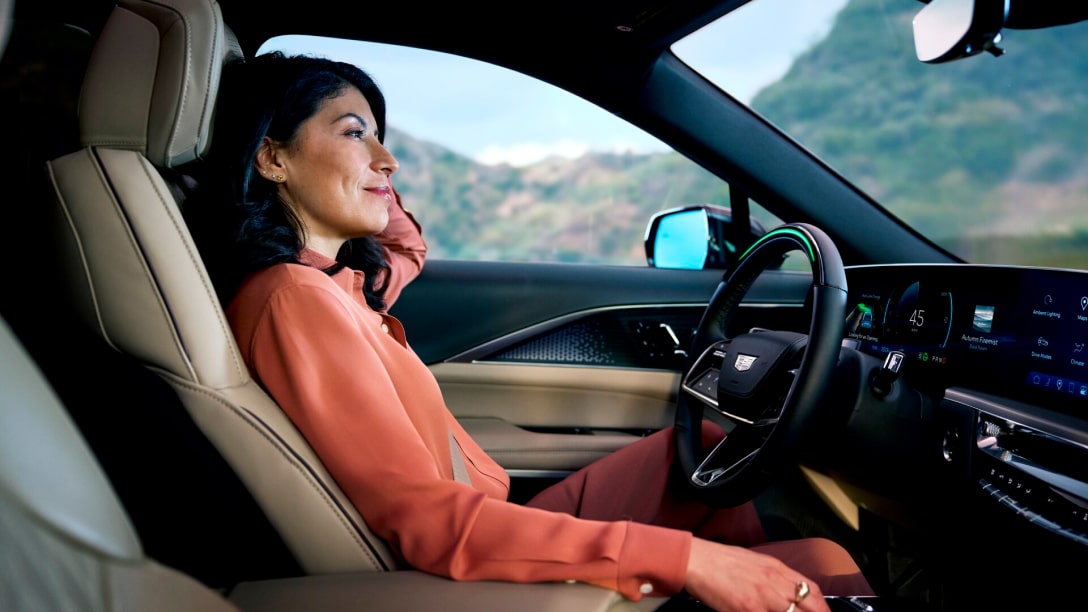 A woman with dark hair, wearing a reddish-orange shirt, is smiling while sitting in the driver seat of a car as she looks out the window