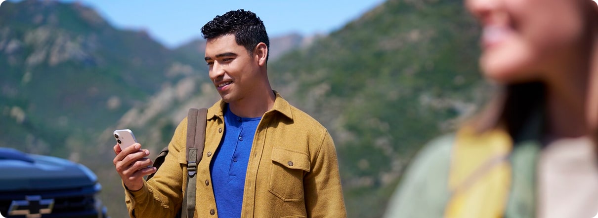 A smiling man with a backpack stands in a rugged, mountainous landscape, looking down at his smartphone, with a vehicle partially visible behind him.