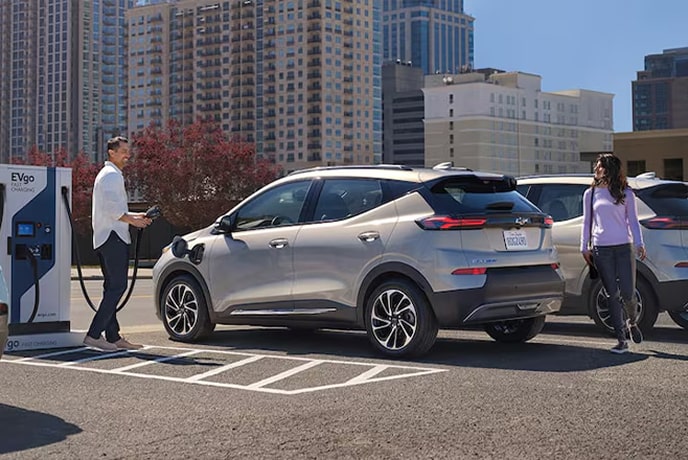 A man charging his EV in a parking lot and a woman walking around the back of the vehicle.