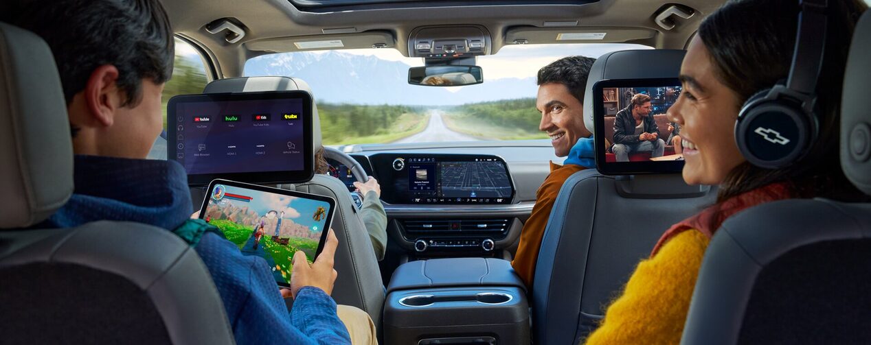 A view from the third row of a vehicle shows a family of four enjoying various forms of entertainment, with two children in the back using a tablet and a mounted screen, while the parents in the front seat are looking at each other and smiling.