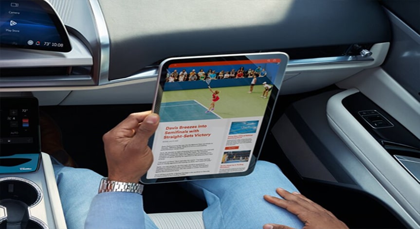 A passenger in the front seat of a vehicle holds a tablet in their lap, using the vehicle wifi, with the screen displaying a live tennis match and a news article about the game.