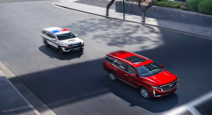 A red Cadillac SUV drives down a city street, being followed by a white police vehicle with its lights flashing.