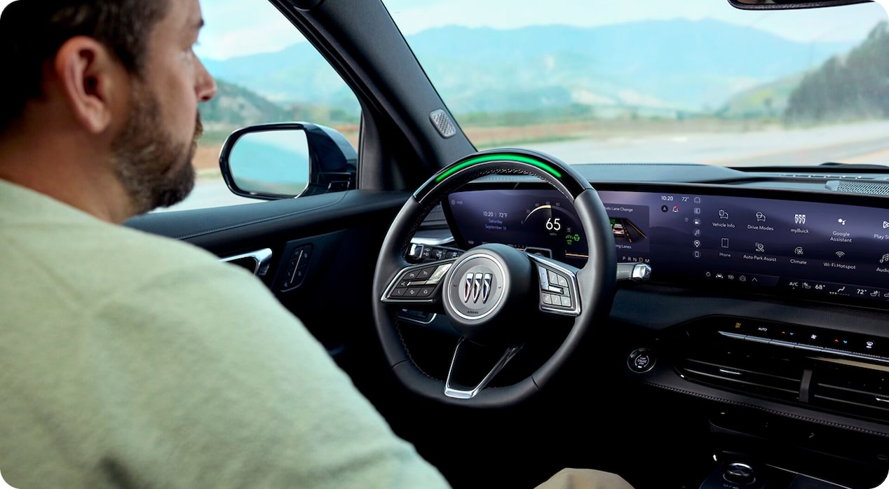 A view from behind a man with a beard, sitting in the driver's seat of a Buick vehicle, with the steering wheel's light bar glowing green to indicate that the hands-free Super Cruise feature is active.