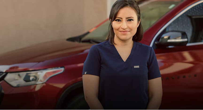 A woman wearing dark blue scrubs stands in front of a red SUV, looking directly at the camera and smiling.