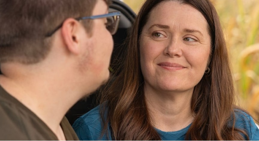 A woman with long brown hair smiles gently while looking at a man, both in an outdoor setting.