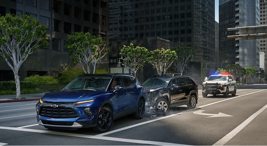 A blue Chevrolet Trailblazer is involved in a multi-car collision on a city street, with a police vehicle approaching the scene.