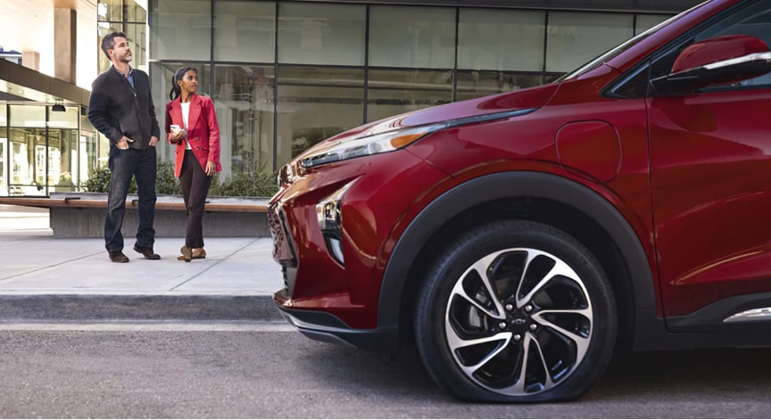 A red Chevrolet Bolt EUV is parked on a city street with two people standing on the sidewalk behind it.