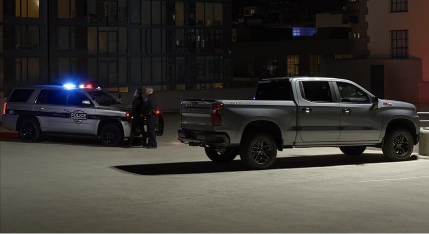 A grey pickup truck is pulled over at night next to a police SUV with an officer standing beside it.