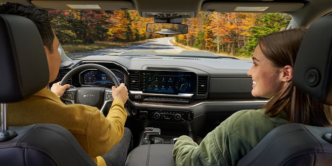 A couple drives a modern Chevrolet pickup truck on a winding autumn road, with a large infotainment screen visible.