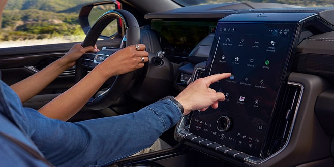 A person's hand touches a large, vertical infotainment screen in a car, displaying multiple app icons.
