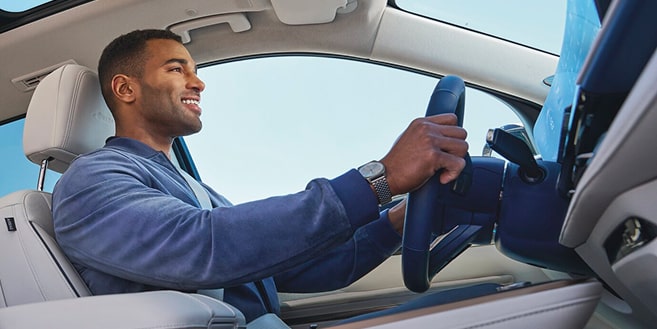 A smiling man in a blue jacket drives a car, with his hands on the steering wheel and a sunroof overhead.