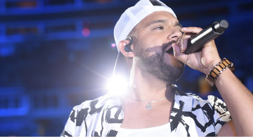 A man in a white cap and patterned shirt sings passionately into a microphone on a brightly lit stage.
