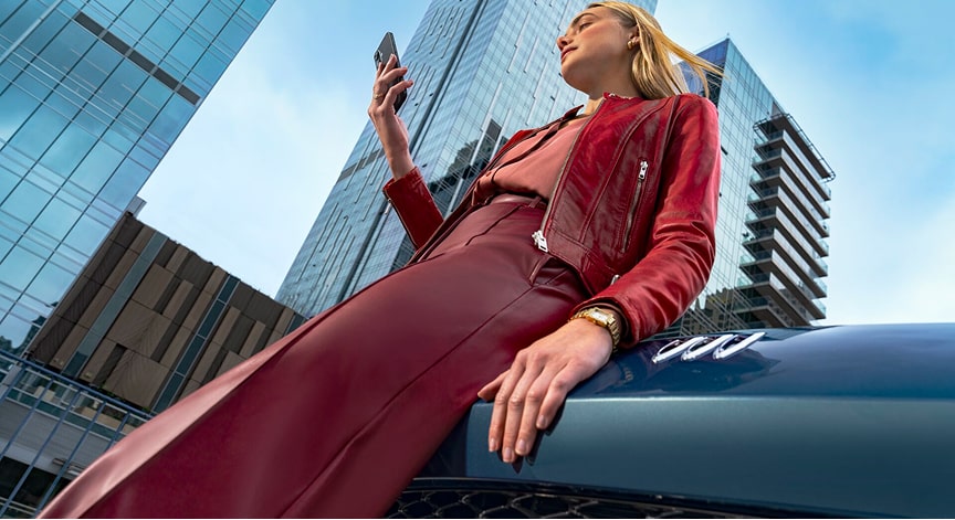 A stylish woman in a red jacket sits on a blue car's hood, looking at her smartphone in front of modern buildings.