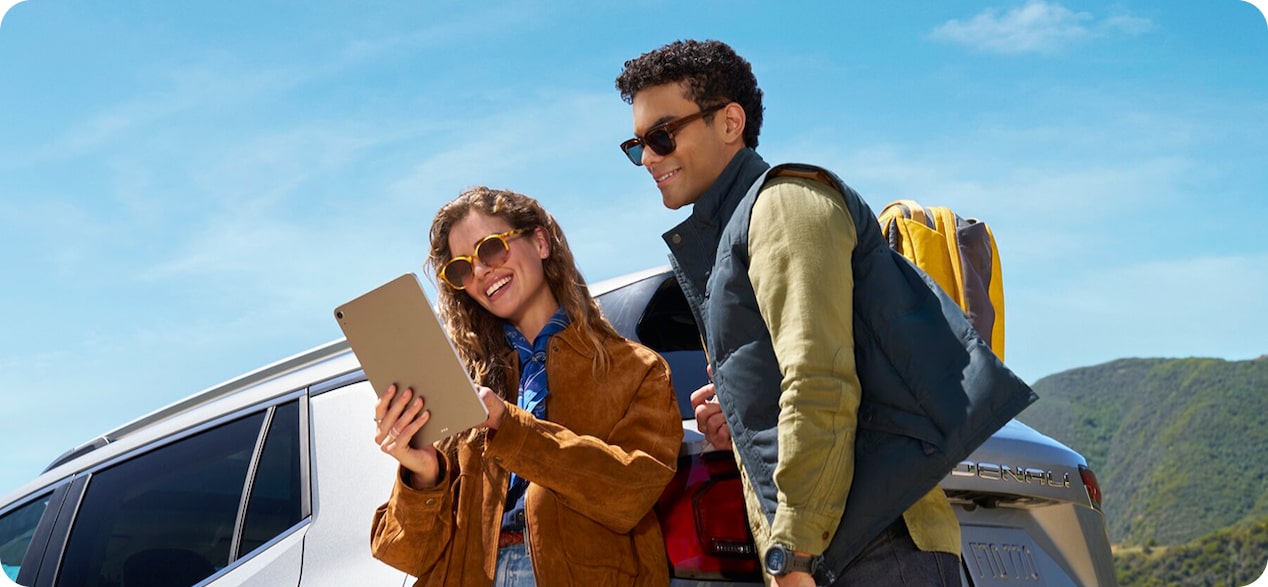 A man and a woman stand by an open car trunk, looking at a tablet with luggage and a mountain backdrop.