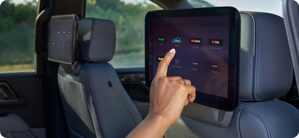 A hand interacts with a rear-seat entertainment screen mounted on a car headrest, showing apps like YouTube and Hulu.