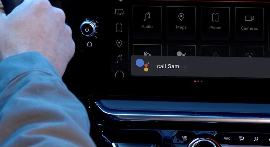 A person's hands on a car steering wheel, with a close-up view of the digital infotainment screen displaying a Google Assistant interface.