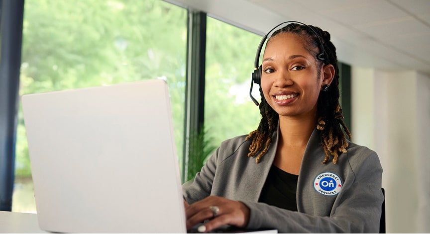 An OnStar customer service representative, wearing a headset, smiling at a laptop with a visible OnStar logo on her jacket.