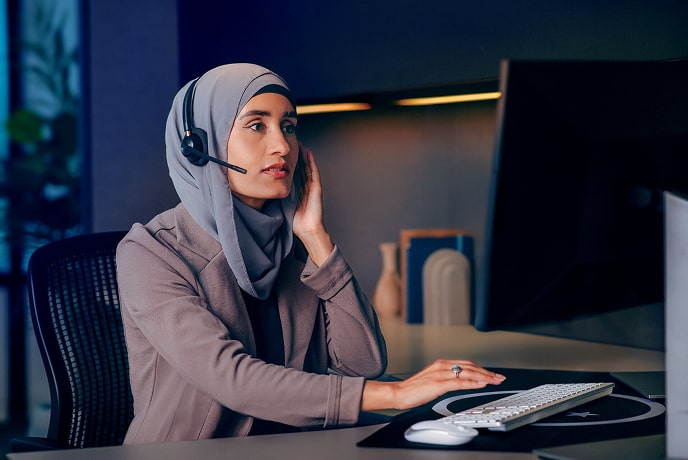 A woman sitting at her desk with her left hand on her headset and her right hand on her keyboard.