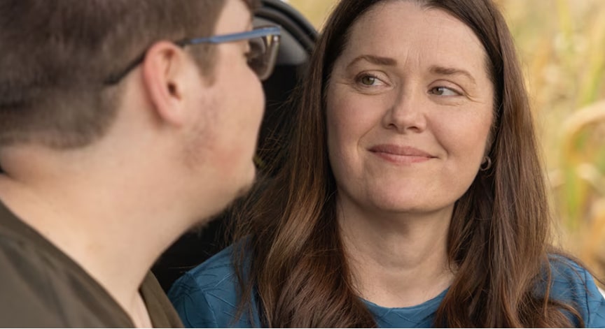 A woman with long brown hair smiles gently while looking at a man, both in an outdoor setting.