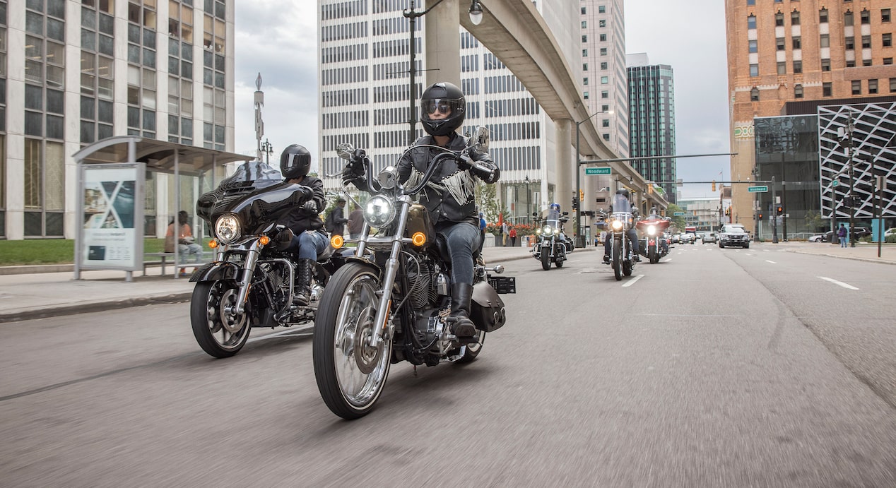 A group of motorcyclists, with riders wearing helmets and jackets, rides down a city street under an elevated track, flanked by tall buildings.