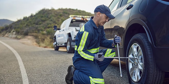 A roadside assistance technician in a blue jumpsuit with neon yellow stripes is kneeling on an asphalt road next to a dark vehicle, using a lug wrench to change a flat tire, with a white tow truck