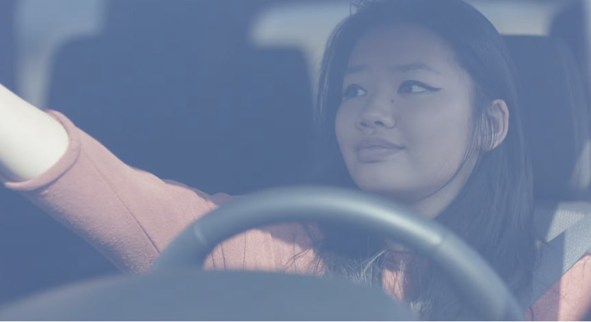 A young woman reaches to adjust the rearview mirror inside a car, with a steering wheel visible through the blue-tinted windshield.