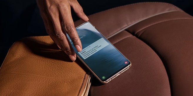 A person's hand reaches for a rose-gold smartphone resting on a brown leather handbag, displaying an OnStar notification that reads "OnStar Emergency Advisor has detected a crash."