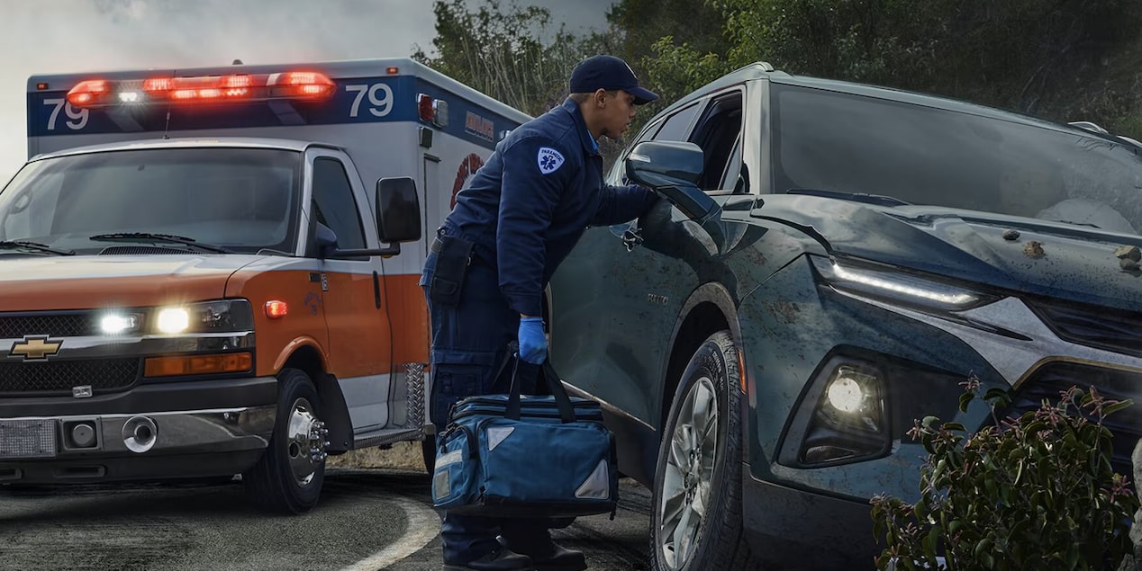 A first responder, wearing a uniform and carrying a medical bag, attends to a damaged dark SUV on the side of a road, with an ambulance with flashing lights visible just behind the vehicle.
