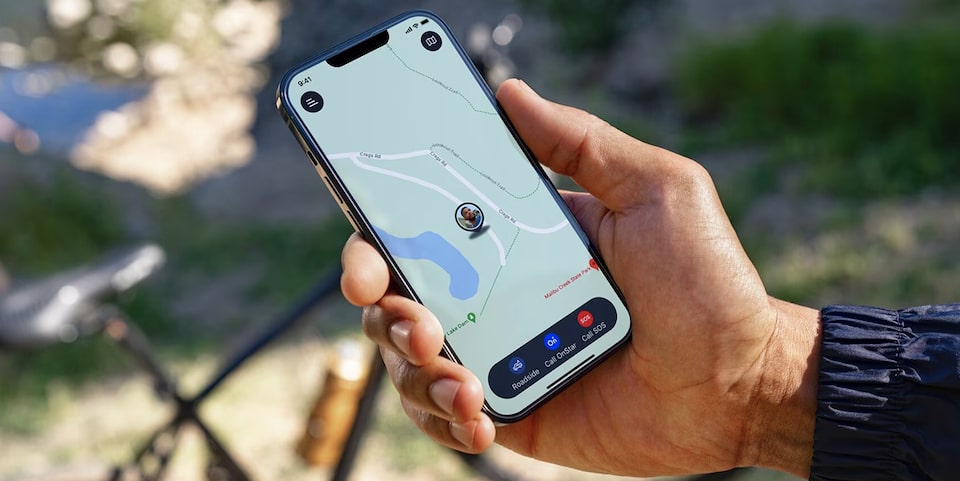 A hand holds a smartphone displaying a map application with the user's location near a lake, showing options to "Roadside," "Call OnStar," and "Call SOS," with a bicycle blurred in the background, suggesting outdoor activity.