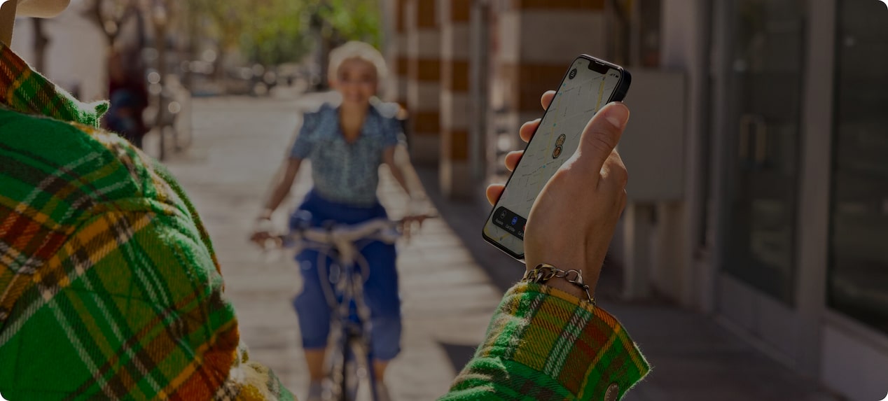 Woman in Distance Riding Bike Towards Man Holding Smartphone