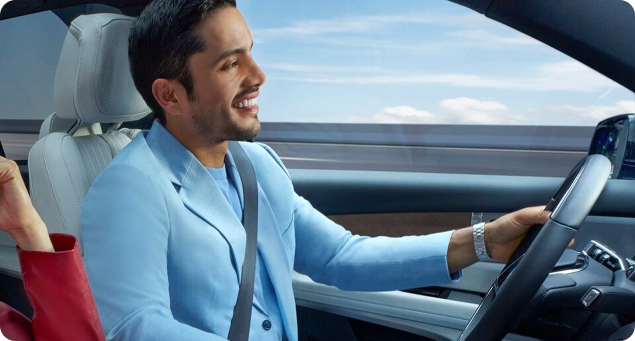 A smiling man wearing a light blue jacket drives a car and looks ahead.