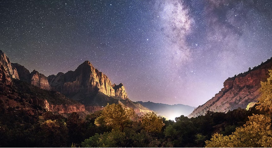 A landscape with stars in the night sky shining brightly over a dark, rugged mountainous landscape.