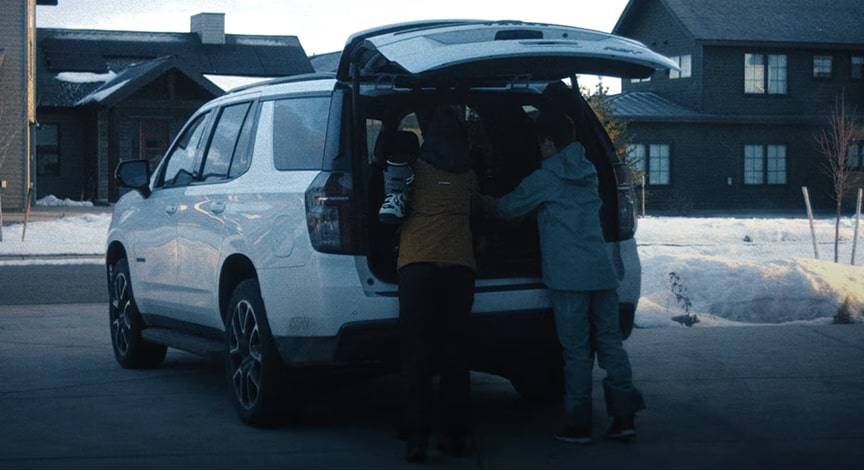 Two people load a white SUV with luggage while the trunk is open on a snowy day.