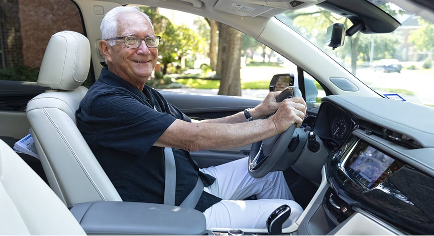A happy older man wearing glasses and a black polo shirt smiles while driving a car.
