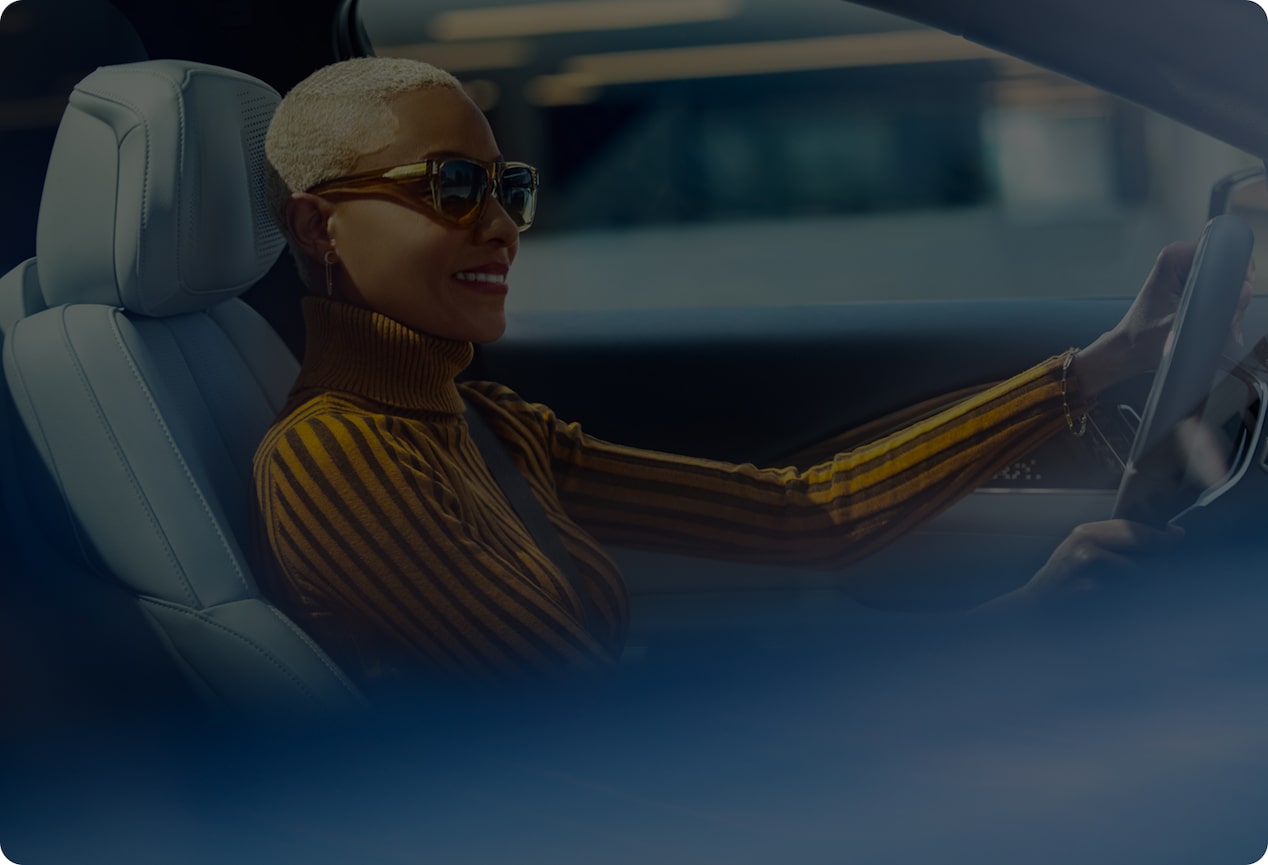 A smiling woman with short blonde hair and sunglasses is driving a car.