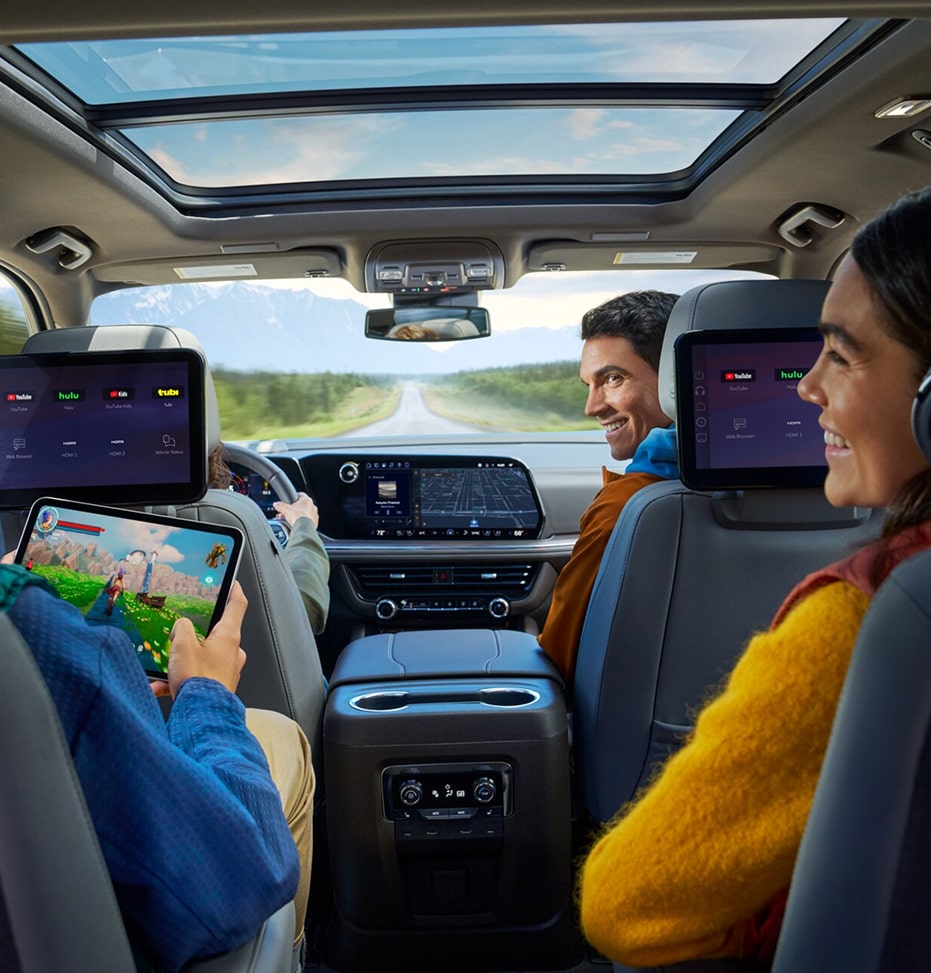 Three people in a car's back seats enjoy entertainment, with one child watching a screen and another playing on a tablet.