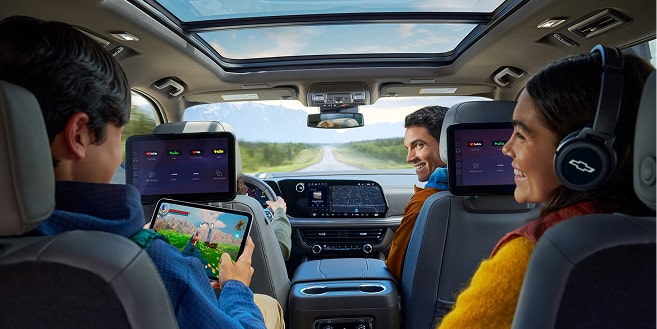 Three people in a car's back seats enjoy entertainment, with one child watching a screen and another playing on a tablet.