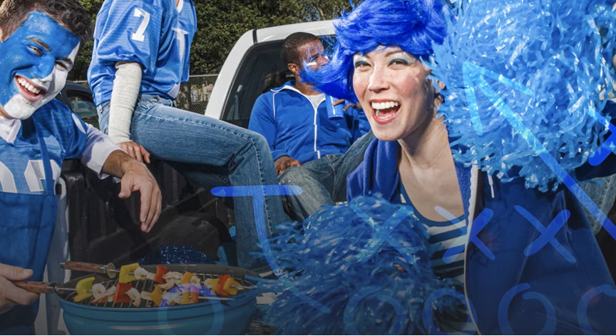Excited people with blue face paint and a woman wearing a blue wig are tailgating at a game.
