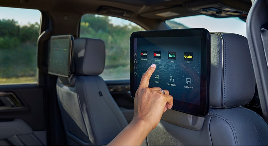 A person's hand touches a touchscreen entertainment display mounted on the back of a car's front seat, showing app icons.