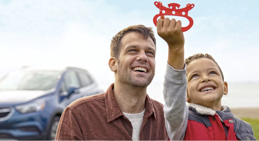 A smiling father and son stand outdoors near a blue SUV, holding up a red toy.